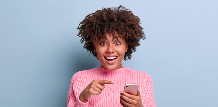 mulher negra com blusa rosa apontando para o celular que está segurando com a mão esquerda e sorrindo