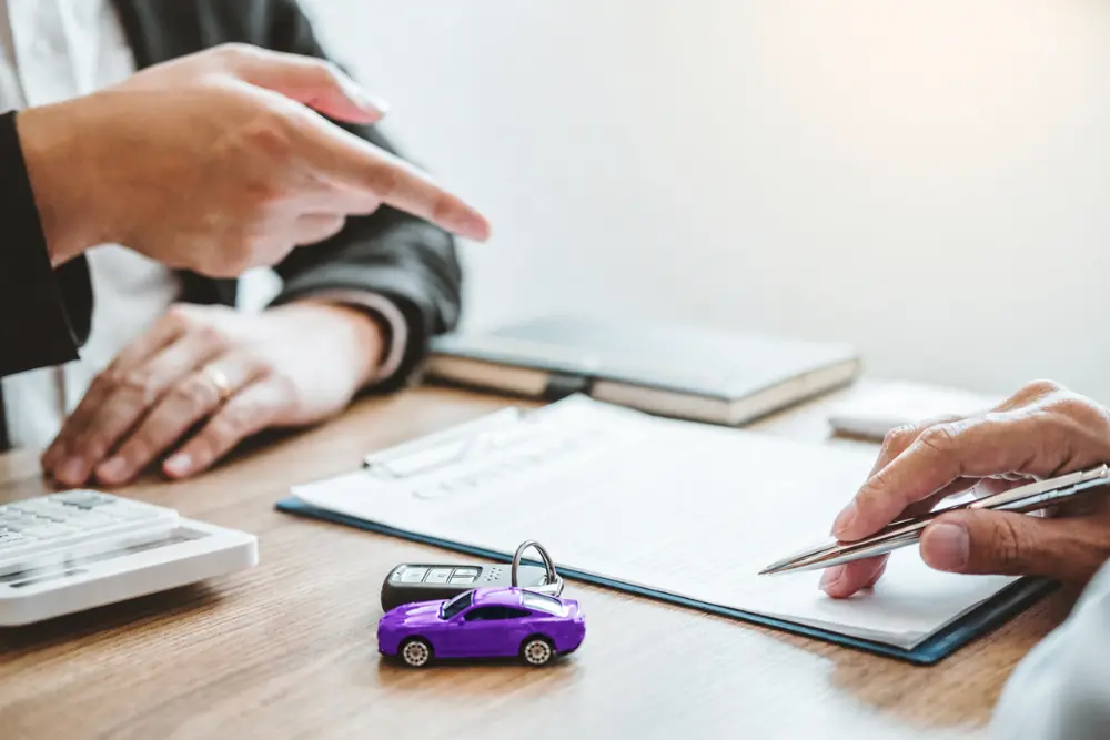 Pessoa assinando contrato com miniatura automotiva representa análise e aprovação de financiamento de carro durante negociação.