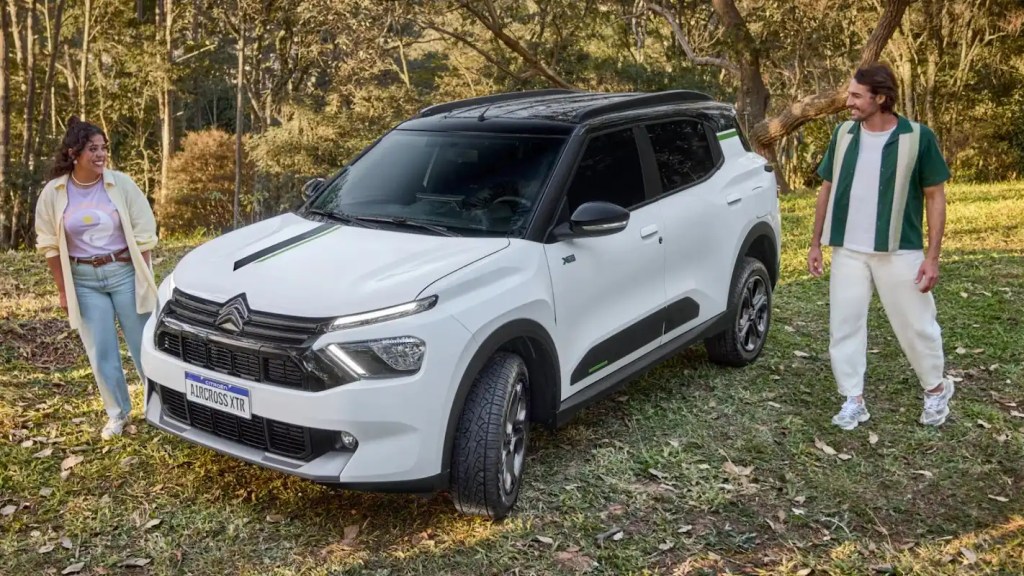 Casal ao lado de um SUV branco estacionado em área verde, com árvores ao redor