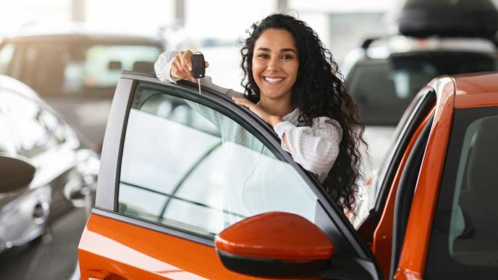 Mulher de cabelos soltos segurando a chave do seu carro, ao lado dele, que é da cor vermelha.