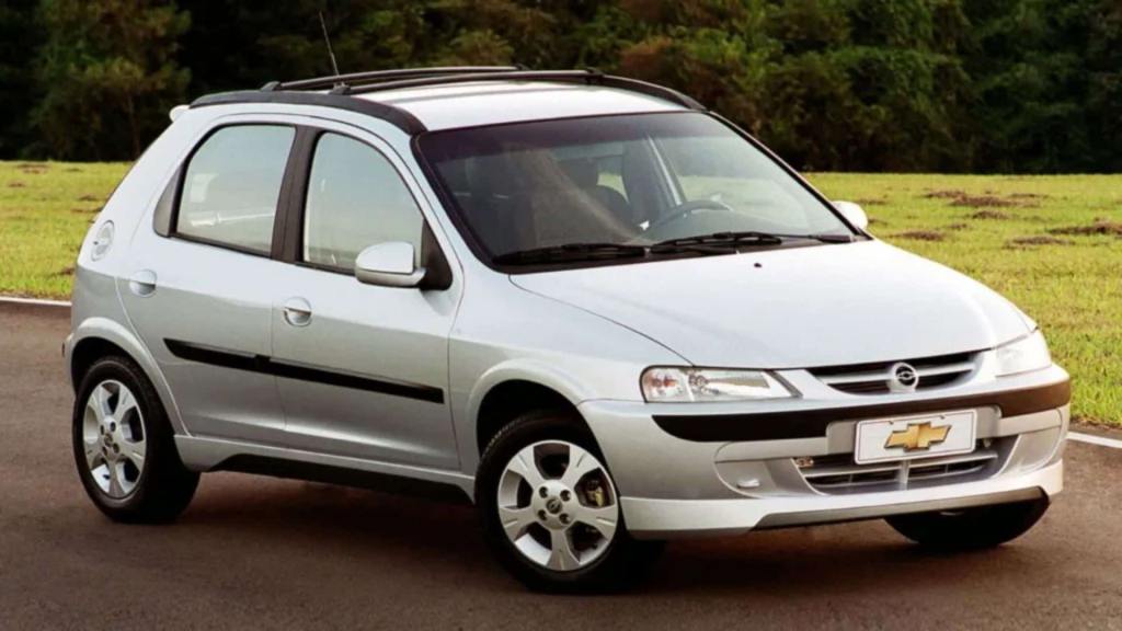 Carro prata do modelo Celta, em estrada com fundo de plantações.