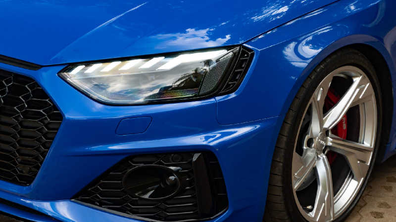 Close-up da frente de um Audi RS4 azul, com faróis LED marcantes, grade esportiva em colmeia e rodas de liga leve com pinças de freio vermelhas, mostrando a performance da Audi mais barato