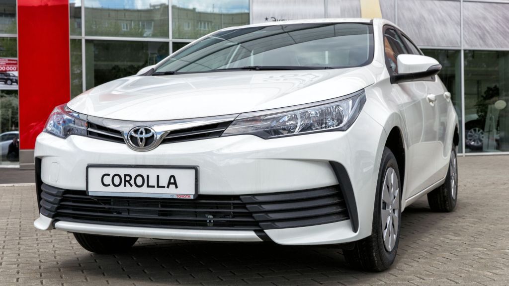 Detalhe frontal de um Toyota Corolla branco reestilizado (versão 2017-2019) em frente a uma concessionária. Entenda quanto custa um Corolla com o facelift.