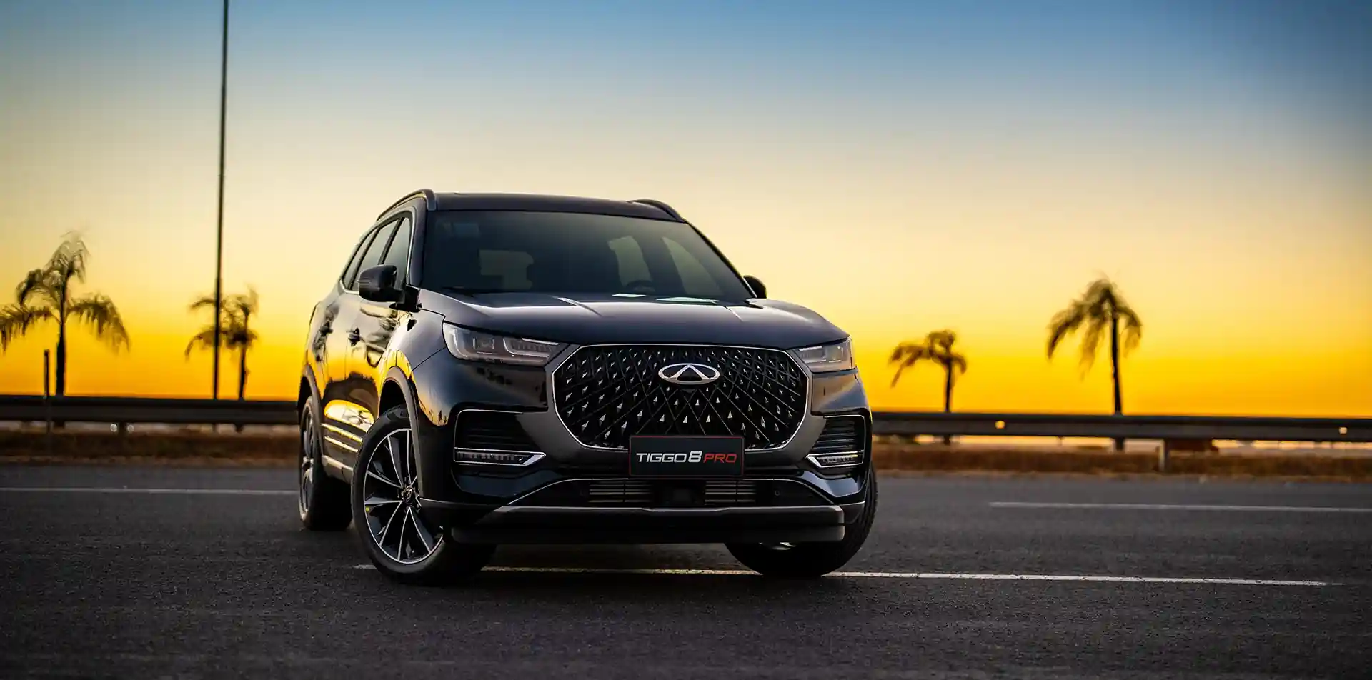 Veículo SUV Tiggo 8 Pro da Caoa Chery, visto de frente em uma rodovia com coqueiros e por do sol amarelado ao fundo