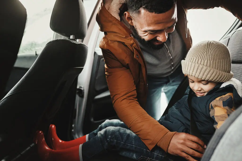 Pai prendendo o cinto de segurança do filho pequeno na cadeirinha no banco traseiro do carro, destacando a importância dos itens de segurança para a proteção da família.